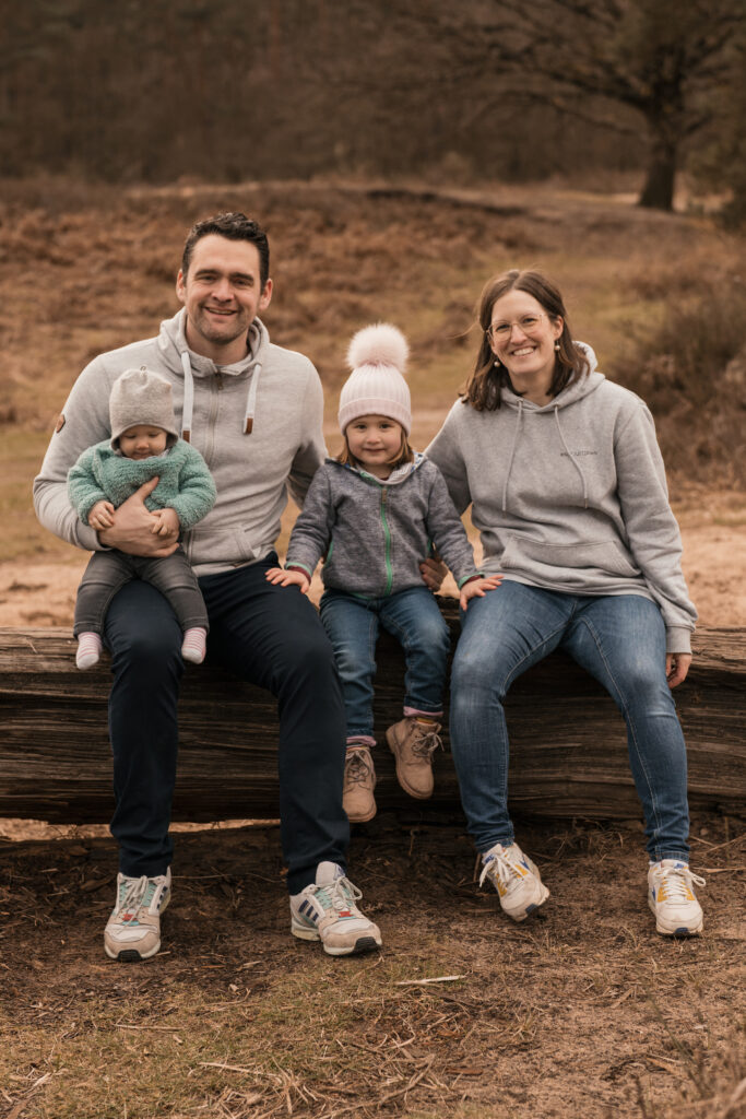 Familie, bestehend aus Mutter, Vater und zwei kleinen Kindern, sitzt lachend auf einem Baumstamm. - Familienfotos Köln