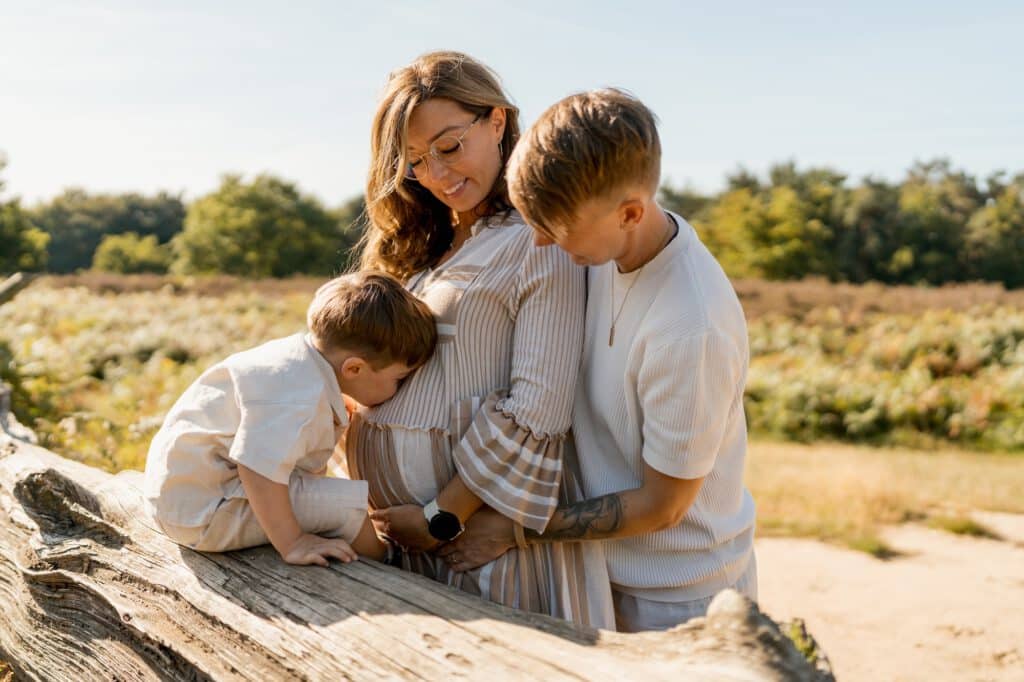 Ein schwangere Frau mit ihren zwei jungen Kindern in der Natur. Das jüngere der Kinder küsst den schwangeren Bauch der Mutter.